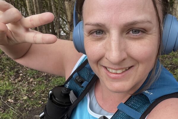 Becci taking a close-up selfie on a wooded path, wearing a sleeveless top with a colourful graphic, over-ear headphones, and a backpack.