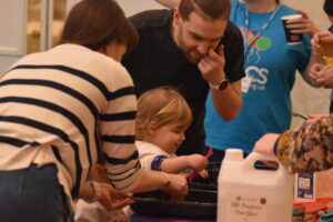 Adults and children at a busy craft table, carefully assembling soft toys and handling materials together."