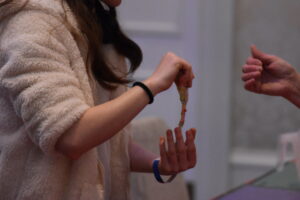 A person holds a small craft item while another person assists, showing a hands-on creative activity."