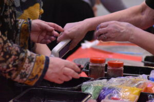 Two people work together on a craft project, using small tools and containers filled with different coloured materials."