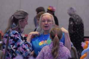 Two people are talking with a MACS staff member, who is engaging with them during the activity session. They are standing together in a busy, friendly craft-room environment."