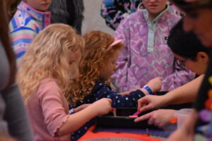 Children gather around a craft table, reaching for materials and concentrating on their creative projects while supported by nearby adults."