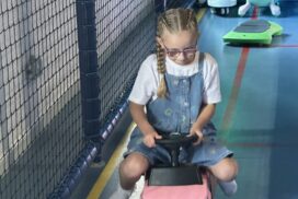 Child wearing glasses sits on a ride-on toy in a soft play area with padded flooring and safety netting."