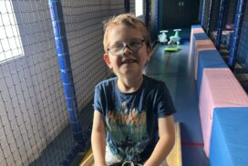 Child wearing glasses seated on a ride-on toy in a soft play area with padded equipment and netted walls."