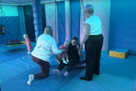 Two adults supporting children as they use a suspended sensory swing in a padded play room with blue lighting."