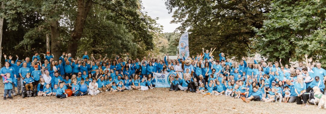 Large MACS community group photo taken outside, with adults and children in blue T-shirts gathered under trees around a MACS banner and Macs the monkey mascot. Many participants are waving at the camera.