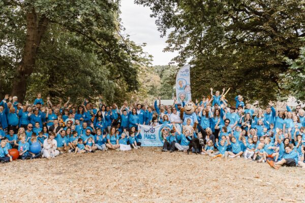 Large MACS community group photo taken outside, with adults and children in blue T-shirts gathered under trees around a MACS banner and Macs the monkey mascot. Many participants are waving at the camera.
