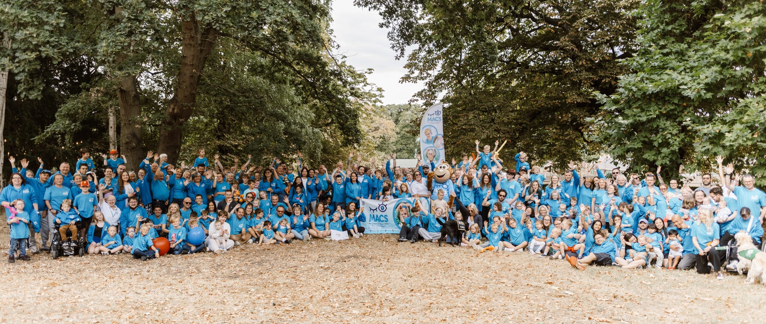 Large MACS community group photo taken outside, with adults and children in blue T-shirts gathered under trees around a MACS banner and Macs the monkey mascot. Many participants are waving at the camera.