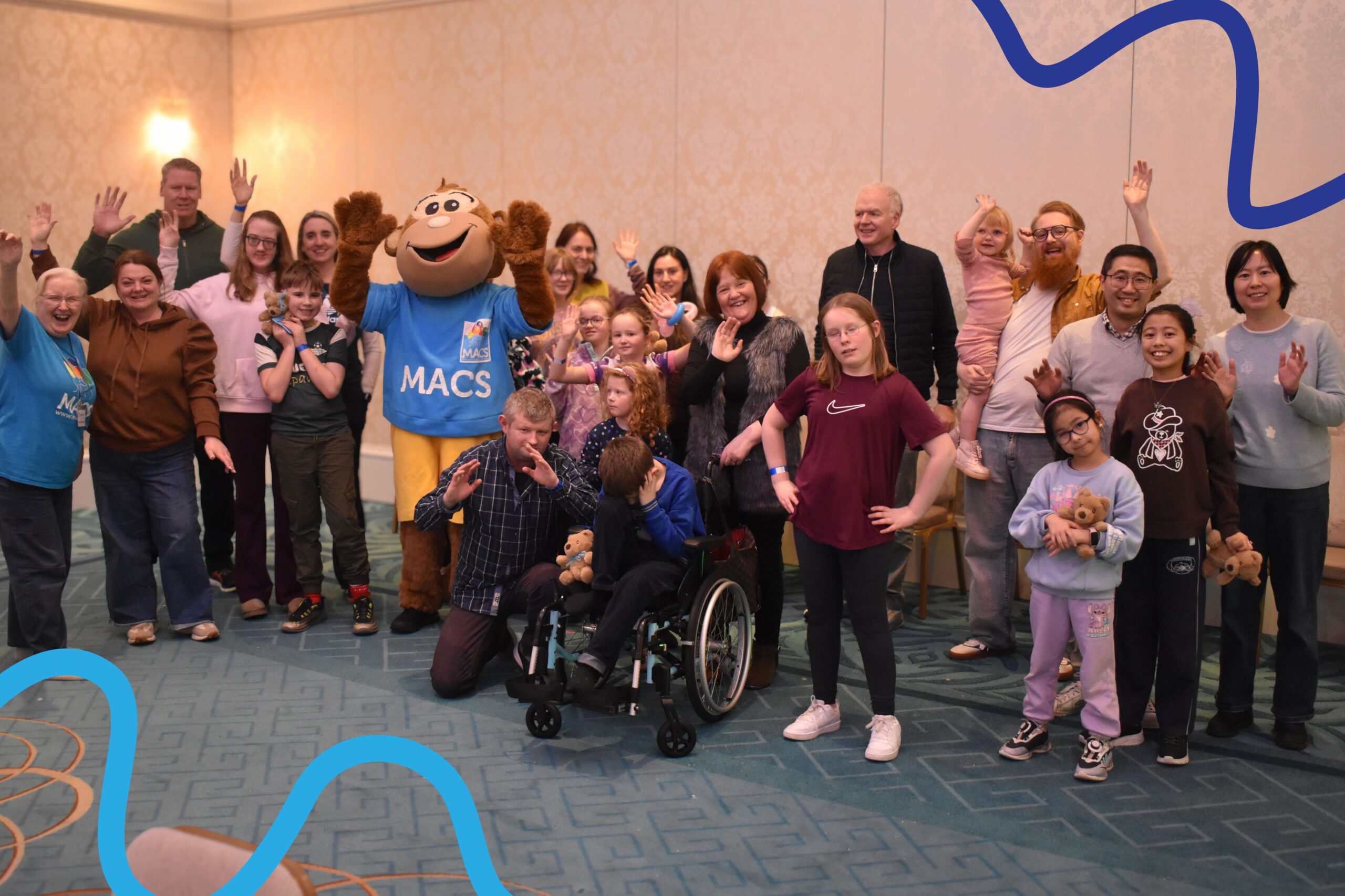 A large group of children and adults stand together in a bright indoor venue with patterned wallpaper and a soft blue carpet. Everyone is gathered around Macs the monkey mascot dressed in a blue MACS T-shirt and yellow shorts. Many of the people in the group are smiling, waving, or posing playfully. One child is sitting in a wheelchair at the front, holding a soft toy, while others stand or kneel around them. Decorative blue graphic lines are added to the corners of the image.