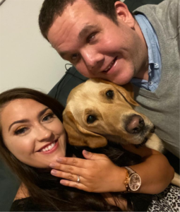 Image of Robbie with Fiancé, Caitlin and dog, Yogi. Caitlin has brown hair and is showing her ring to the camera. Robbie is on the right-hand side and has brown short hair. Yogi is a Golden Lab Retriever Guide Dog and sits between Caitlin and Robbie.