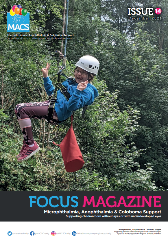 This cover features a person participating in an outdoor zip‑lining/high‑ropes activity. The person is suspended in midair, wearing safety gear including a helmet, harness, and rope attachments. They are surrounded by tall, dense green trees, indicating a forest setting. The top displays the MACS logo and “ISSUE 14.” The bottom banner carries FOCUS MAGAZINE with the subtitle Microphthalmia, Anophthalmia & Coloboma Support.
