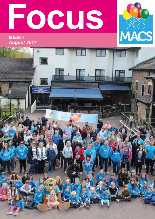 The cover shows a large outdoor group photo taken in front of several buildings with white and cream façades. A crowd of people is gathered, many wearing bright blue clothing, such as jackets or T‑shirts, forming a unified group. Some people are seated on the ground, while others stand behind them. The scene appears to be part of an organised community event. The top banner is bright pink with the title “Focus”, the text “Issue 7 Summer 2017”, and the MACS logo featuring colourful balloons.
