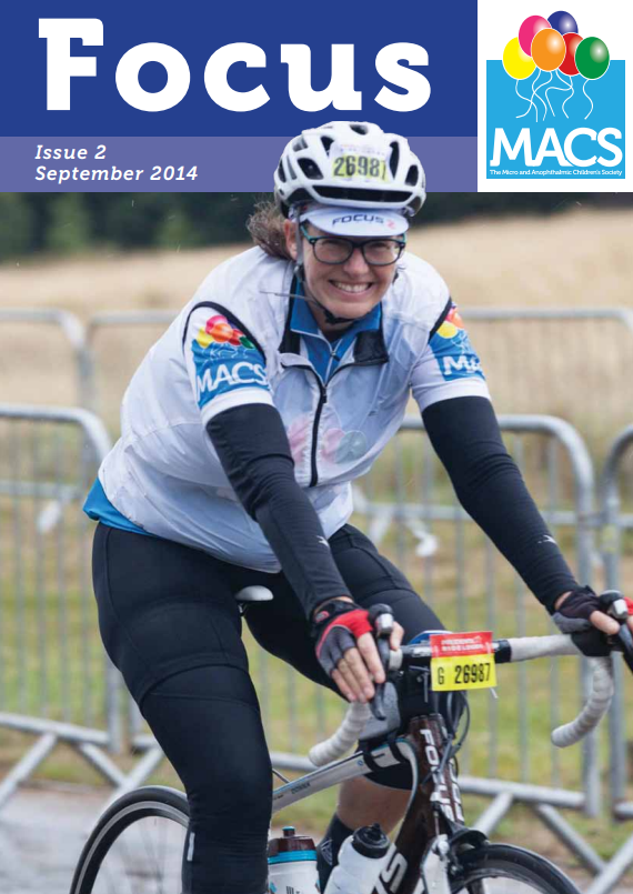 The cover features a person participating in a cycling event. The person is wearing a white and blue cycling jacket with dark sleeves, black cycling shorts, gloves, and a helmet with sunglasses. A race number is attached to the bicycle’s handlebars. The individual is positioned on a road with metal barriers in the background, suggesting an organised outdoor event. The top section of the cover has a blue banner with the title “Focus”, the text “Issue 2 September 2014”, and the MACS logo with colourful balloons.